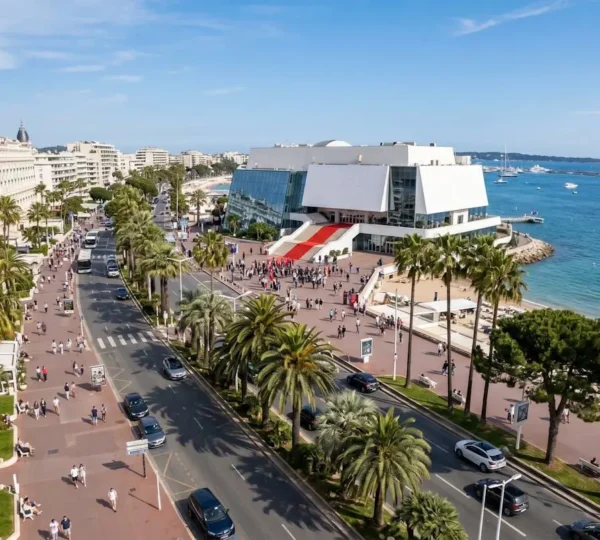Vue panoramique de la Croisette à Cannes avec le Palais des Festivals et ses escaliers rouges en arrière-plan, bordée de palmiers et de la mer Méditerranée